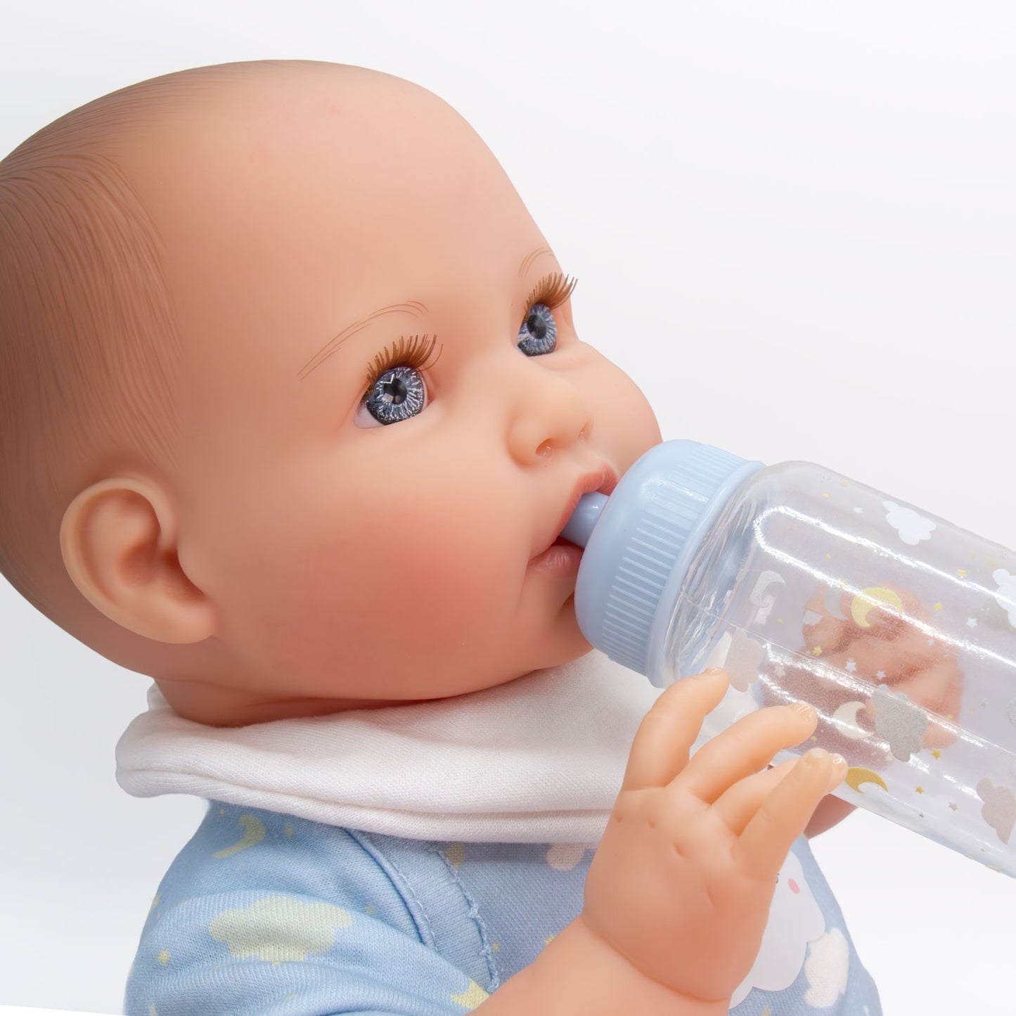 A realistic baby doll with blue eyes and a pacifier clip, wearing a light blue cloud-patterned outfit and white bib, drinking from a clear bottle with a light blue cap.