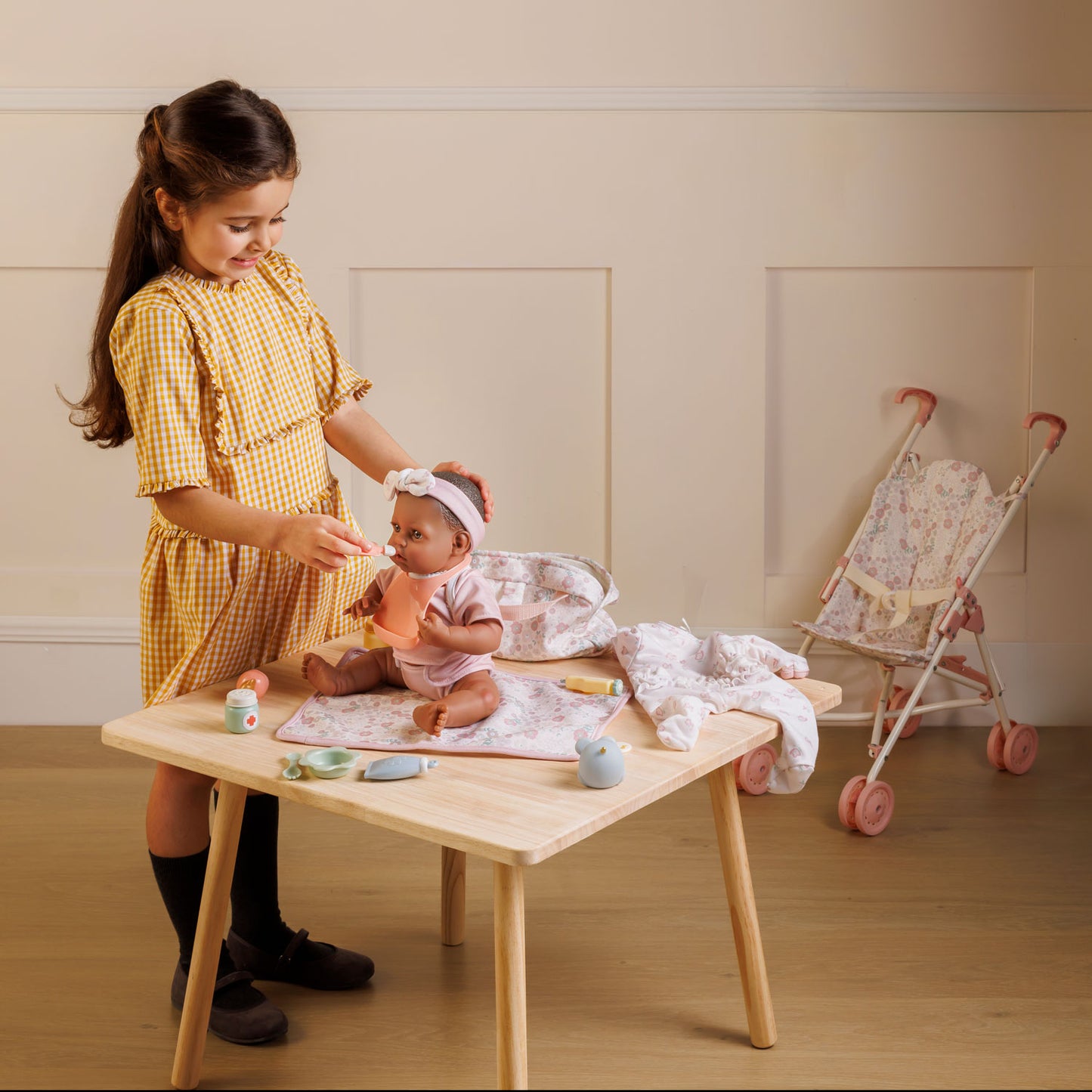 Child playing with a baby doll at a wooden table in a room with a crib in the background.