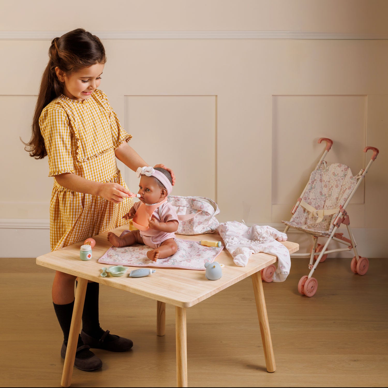 Child playing with a baby doll at a wooden table in a room with a crib in the background.