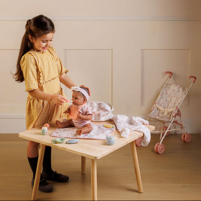 Child playing with a baby doll at a wooden table in a room with a crib in the background.