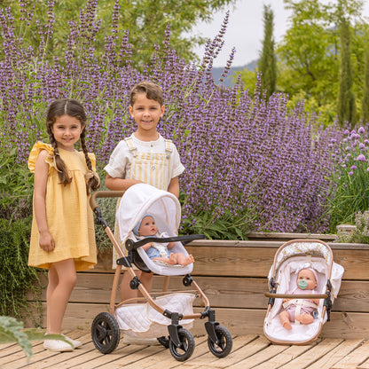 A baby stroller and matching bassinet are shown outdoors on a wooden deck in front of a garden of purple flowers, with two children standing beside them. The stroller has a white canopy and cream quilted interior with a rose-gold frame, and the bassinet is placed to the right.