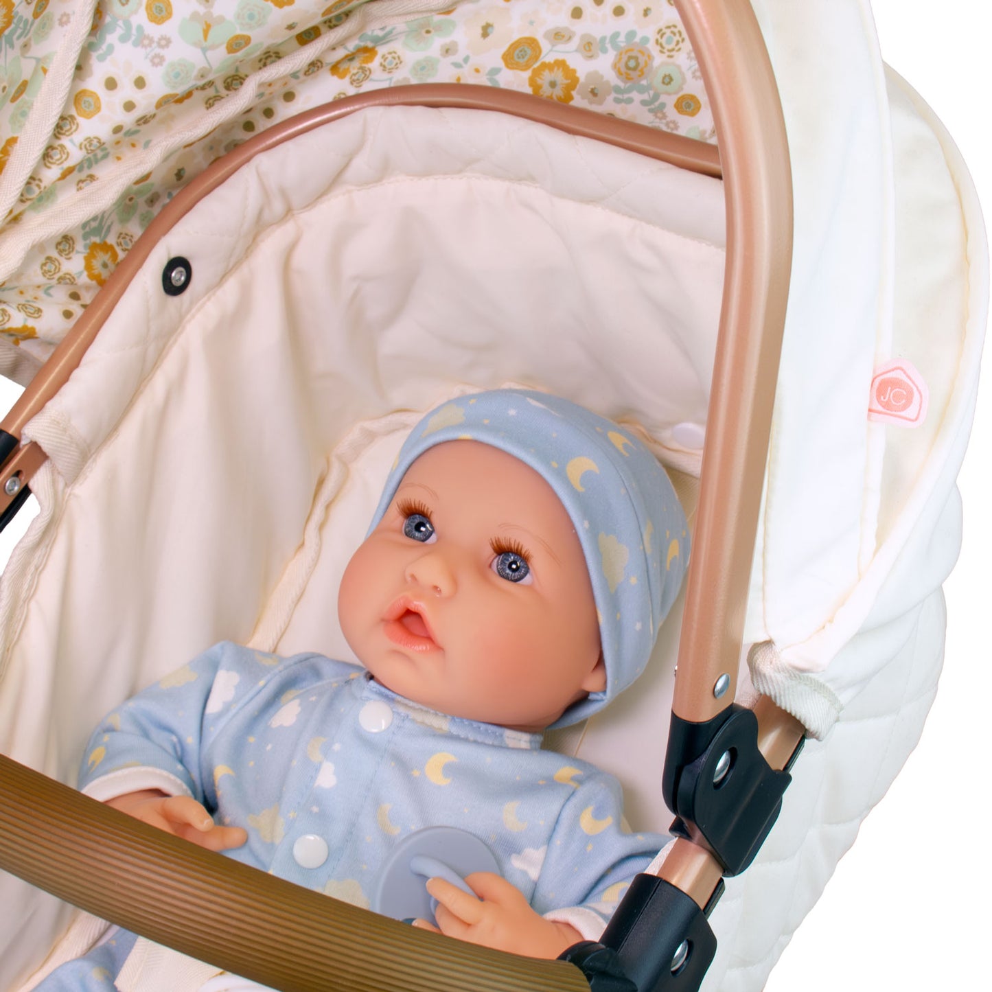 A realistic baby doll with blue eyes wearing a light blue moon and star patterned onesie and matching bonnet, lying inside a white baby stroller with a rose gold frame.