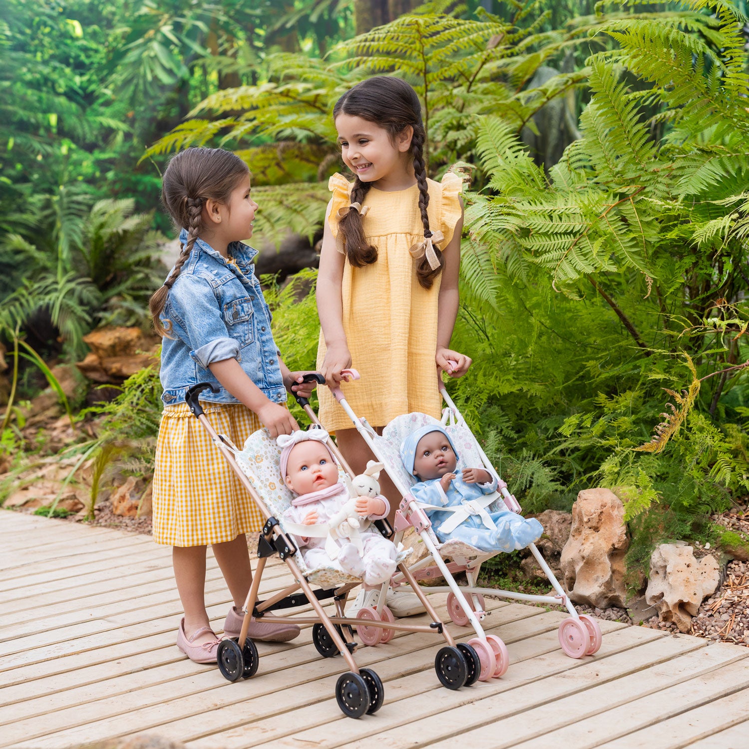 Two young girls with dolls in a stroller outdoors among greenery