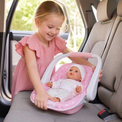 Smiling girl places 12.5" Nonis baby doll in a pink polka dot carrier on a car seat.
