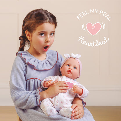 A young girl with braided brown hair sits indoors holding a realistic baby doll. The girl wears a light blue ruffled long-sleeve dress, and the doll is dressed in a white outfit with pink floral patterns and a pink bow headband.