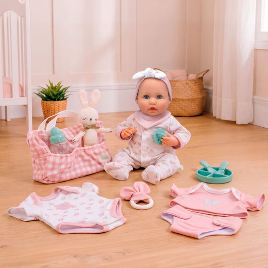 A doll dressed as a baby sits on a wooden floor surrounded by baby clothing, a pink gingham basket with a bottle and plush bunny, and small accessories in a softly lit nursery-like room.