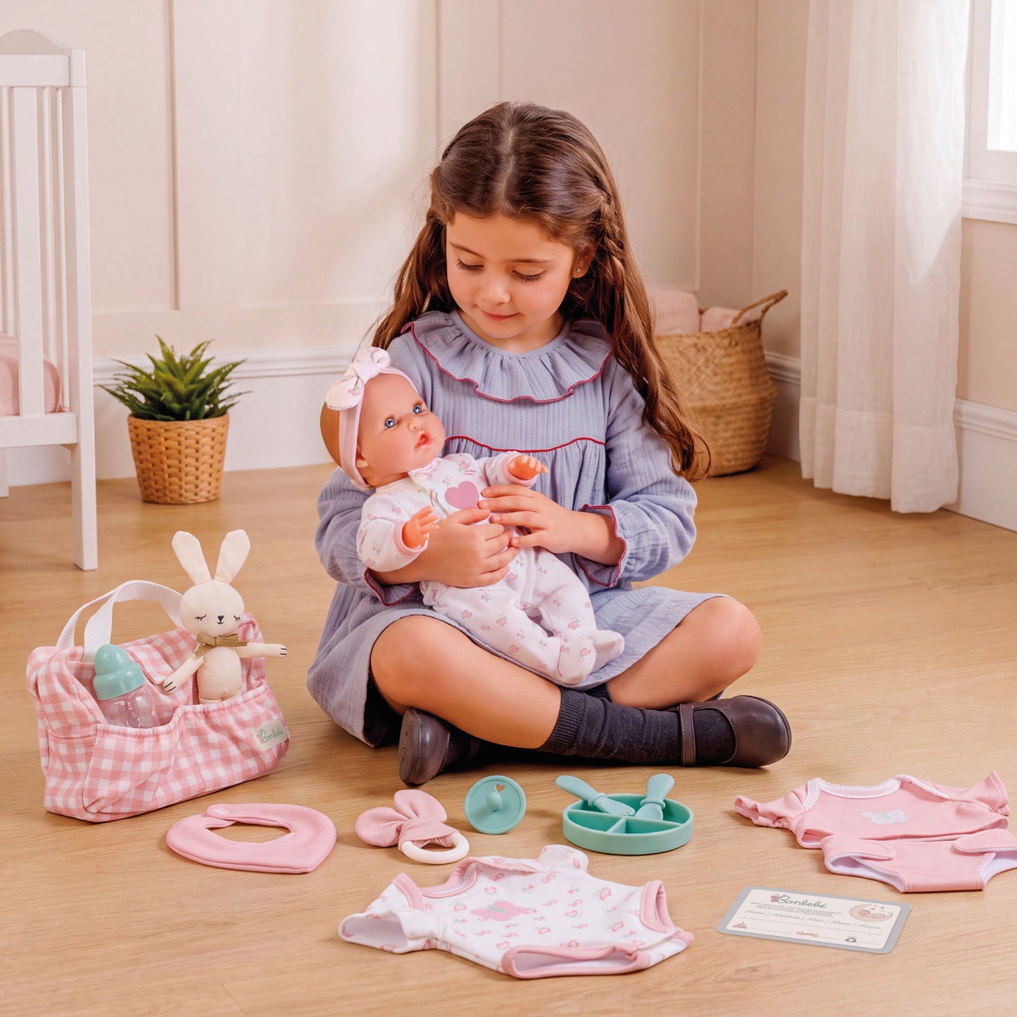 A young girl sits on a wooden floor holding a baby doll, surrounded by a pink and white baby play set with a gingham bunny bag, small accessories, and clothing pieces.