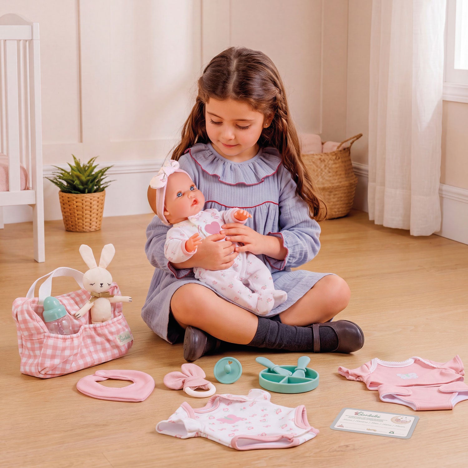 A young girl sits on a wooden floor holding a baby doll, surrounded by a pink and white baby play set with a gingham bunny bag, small accessories, and clothing pieces.