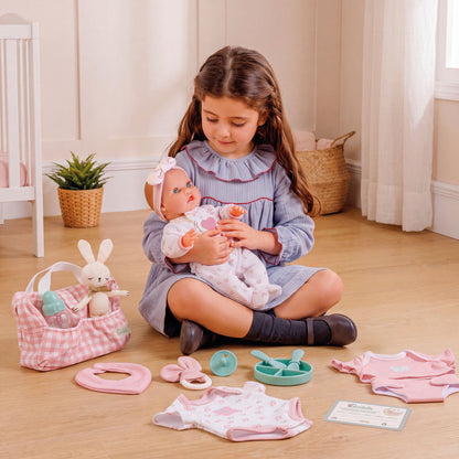 A young girl sits on a wooden floor holding a baby doll, surrounded by a pink and white baby play set with a gingham bunny bag, small accessories, and clothing pieces.