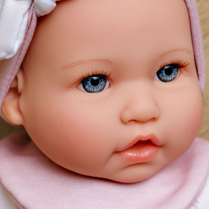 Close-up of a realistic baby doll or toy baby with large blue eyes, pink skin tones, a pink bonnet with a white bow, and a pink bib.